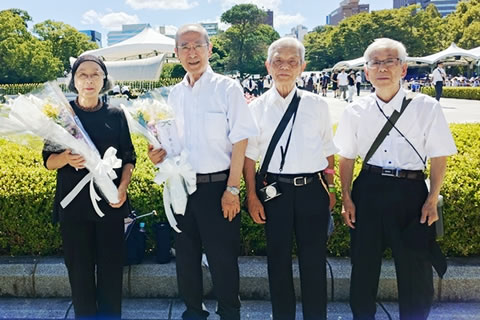 広島平和公園内で横並びに立つ4人の集合写真。背景奥に式典のテントや死没者慰霊碑の屋根などが見える。