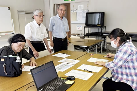 向かい合わせの長机に、広島市職員1名と東友会代表1名が向き合って座り、それぞれの手元の書類を見ながら話をしている。東友会代表2名が脇に立って書類を見ている。