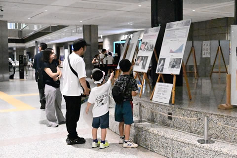 大人が2人の子どもとともに立ち止まり、パネルを見ている。小さい子供はパネルを指さしている。パネルは高さ1メートルほどあるように見え、ひな壇のような土台とイーゼルの高さを合わせ、上端は大人の身長より高い位置にある。