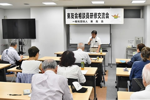「地区相談員研修交流会」と大きく横書きされた紙が、会場前方のホワイトボードに張られている。その前のに講師が立って、マイクを使って話している。並べられた机に着席した参加者が資料に目を落とすなどしながら聞いている。