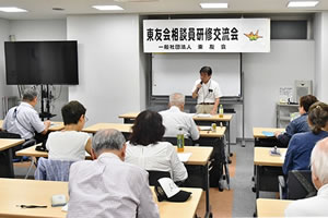 「地区相談員研修交流会」と大きく横書きされた紙が、会場前方のホワイトボードに張られている。その前のに講師が立って、マイクを使って話している。並べられた机に着席した参加者が資料に目を落とすなどしながら聞いている。