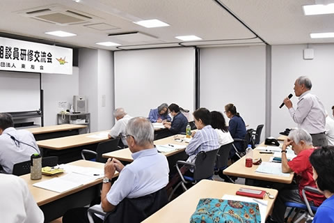 参加者たちのうち1人が席で立ち、マイクを使って放している。他の参加者たちは着席して聞いている。