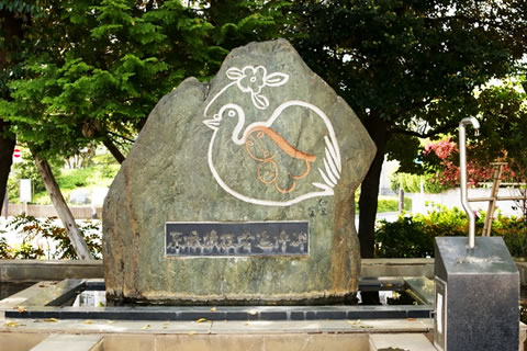 5枚の花弁のある花と3枚の葉が先端にある枝をくわえた鳩のなかに、女性が描かれている絵が彫られている追悼碑