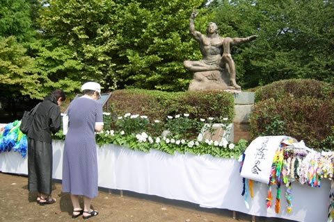 三鷹市平和の像 8月15日「平和のつどい」にて。像の手前に献花台がしつらえてあり、花や千羽鶴がおかれている。二人の人が像に向かって手を合わせて立っている。