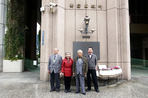 「新宿区役所」正面入り口の柱に設置された銘板の前に、4人の人が立っている。