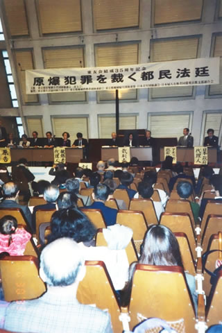「原爆犯罪を裁く都民法廷」と書かれた横幕のかかる舞台の上に机が並べられ、「原告」「被爆者代理人」「アメリカ政府代理人」「日本政府代理人」「裁判長」が座っている。会場の座席は参加者で埋まっている。
