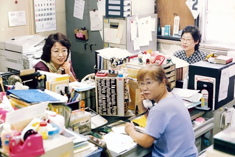 書類や書類棚、スタンプのケースなど、あふれるほど物が積まれた事務机が接して置かれ、事務局員3人がそれぞれ座って電話応対など仕事をしている。