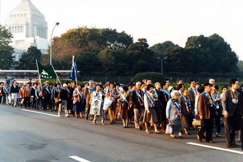 たすきを掛け、旗を持つなどして歩く被爆者たち。背景に国会議事堂が写っている。
